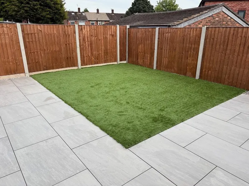 Neat rectangular artificial grass lawn bordered by large light-grey patio tiles, enclosed by a stained wooden panel fence with concrete posts.