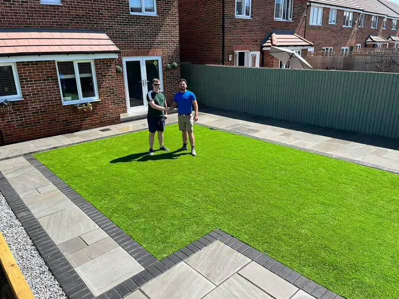 Fresh artificial grass lawn laid next to a natural stone patio at the rear of a red brick house, with a dark block-paver edge and vertical-slat timber fencing.