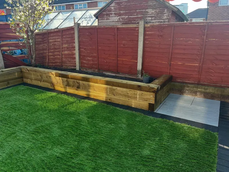 Vibrant artificial turf beside an L-shaped timber sleeper retaining wall, with grey paving and a wooden lap-panel fence in the background.