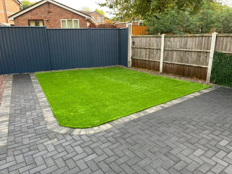 Artificial grass installation framed by grey herringbone block paving and a buff-toned border, set between modern blue-grey fencing and traditional wooden panels.
