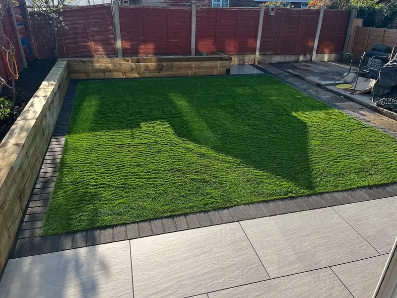 Striped artificial grass bordered by charcoal block pavers and light patio slabs, with new timber sleeper raised beds along a wooden fence.