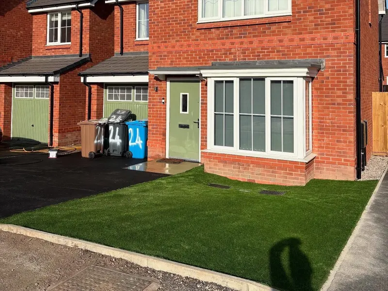 Small front artificial grass area separated from a smooth black tarmac driveway by a concrete kerb, in front of a red brick house with a green door.