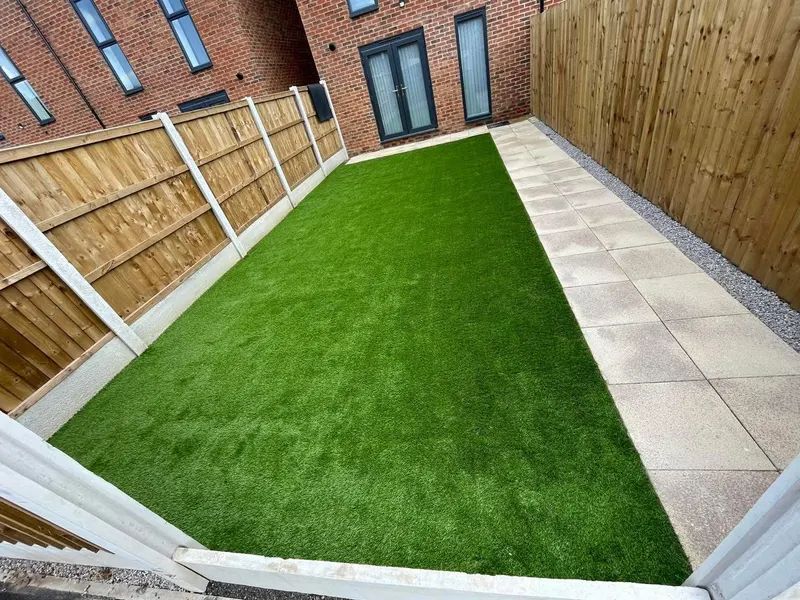 Level artificial grass lawn running alongside a row of light square paving slabs and gravel, bounded by vertical-slat timber fencing and a brick house.