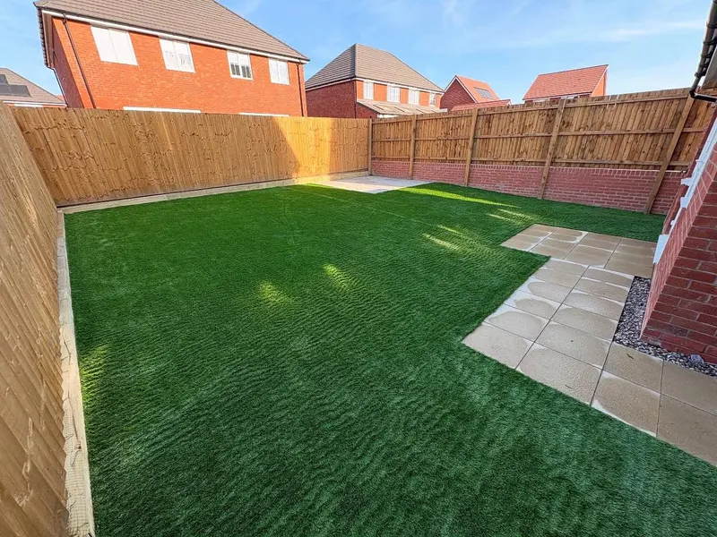 Large artificial grass area enclosed by wooden fencing on a low red brick wall, with a small light stone patio at the house corner.