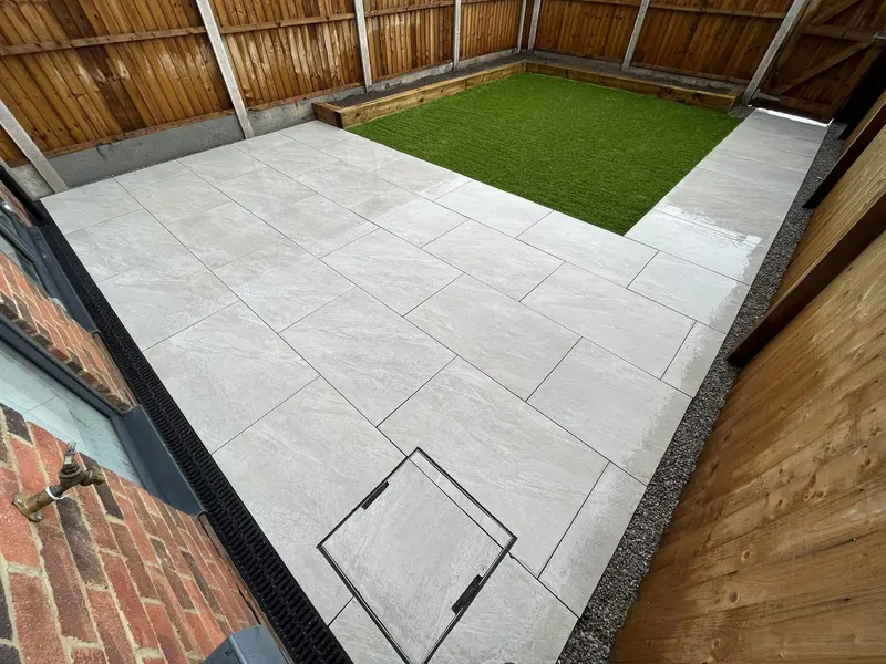 L-shaped light-grey patio slabs surrounding a square artificial grass lawn edged with timber sleepers, against a vertical-slat fence and brick wall.