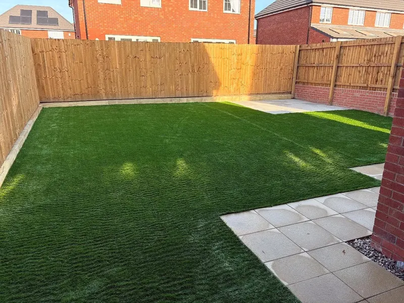 Artificial turf laid in an L-shape beside cream square paving slabs, with timber slat fencing mounted on a red brick garden wall.
