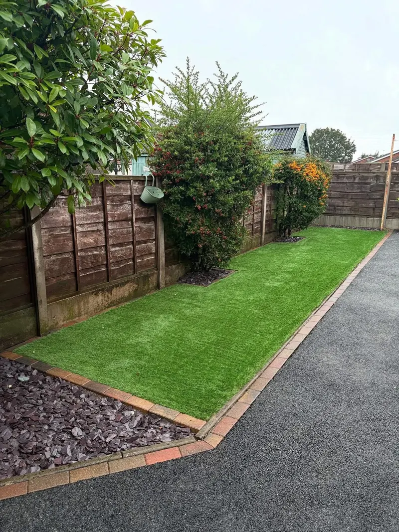 Artificial grass lawn with brick-paver edging, shrub plantings along a wooden fence, and a smooth tarmac strip separated by the same edging.