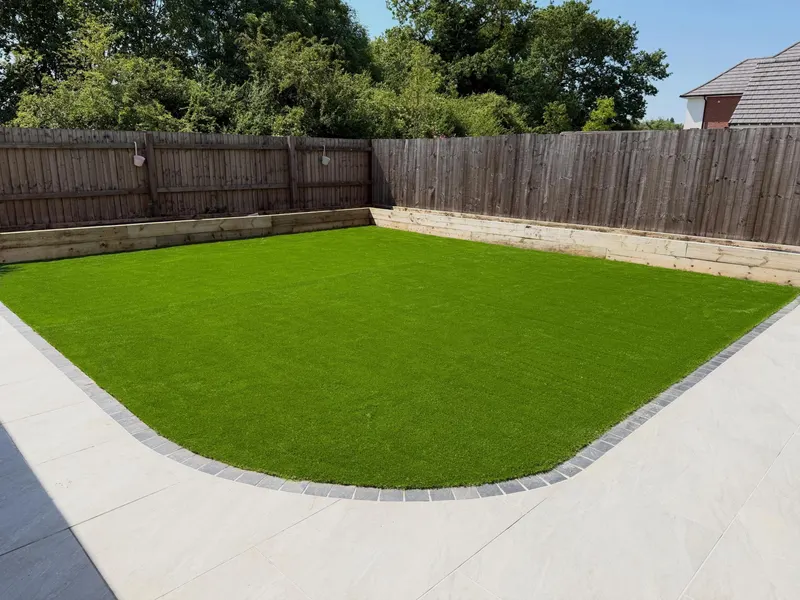 Artificial grass with a curved front edge, dark cobble sett border, light square patio tiles, and double-tier timber sleeper raised beds against slat fencing.