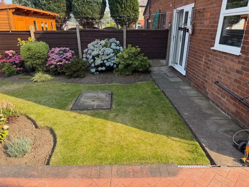 Sunny back garden with a green lawn area, pink hydrangeas and shrubs along a wooden fence, paved path beside a red brick house, and a utility cover in the grass.
