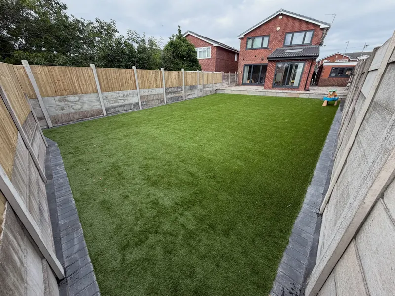 Large rectangular artificial grass lawn edged with dark block pavers, opening from a light stone patio with bifold doors on a red brick house.
