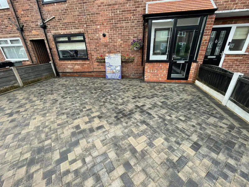 Wide charcoal grey block driveway laid herringbone, edged with a light tan soldier border that continues into a side access strip in front of a red brick semi-detached house.