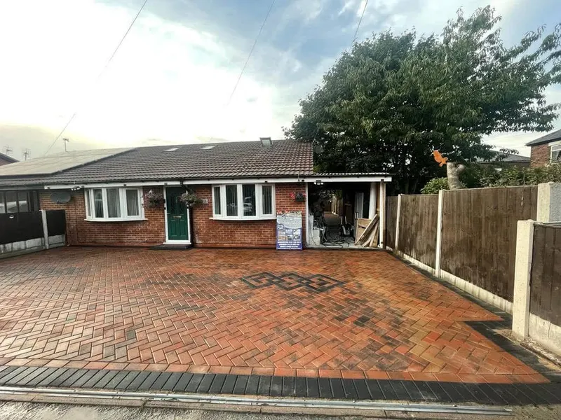 Dark charcoal herringbone paving from the street past a bay window, framed by grey and charcoal border courses that narrow into a side path toward a garage.