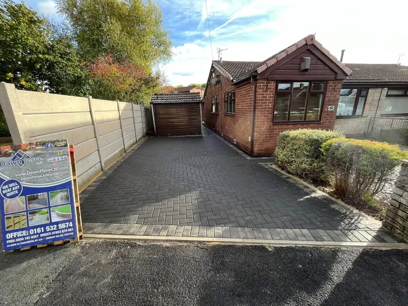 Large rear garden surfaced in charcoal herringbone blocks with a double charcoal border, curving past a small lawn, gravel area and a raised porch on brick pillars.