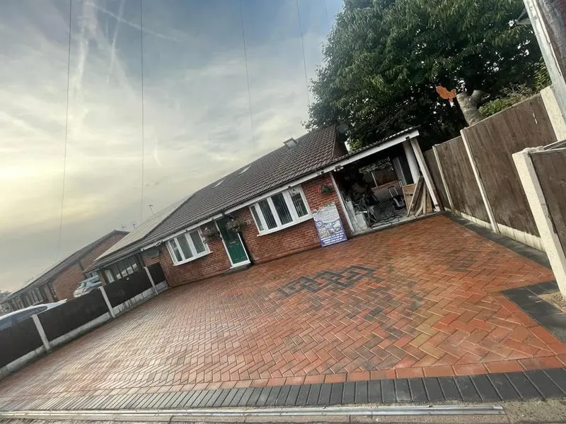 Long charcoal herringbone driveway between a concrete panel fence and red brick bungalow shrub bed, with lighter grey block edging at the entrance and along the sides.