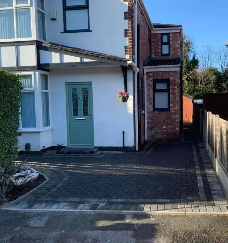 Multi-tonal red and brown herringbone paving filling a front forecourt and carport area, framed in charcoal blocks with a decorative interlocking diamond feature in the centre.