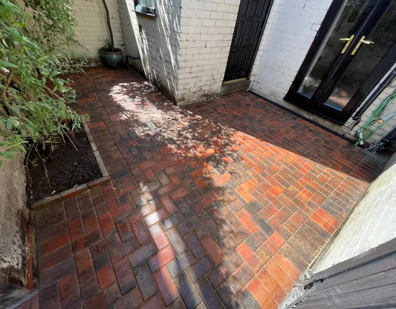 Red and orange herringbone block forecourt with a double charcoal border following a sweeping curve, with a manhole cover set flush in the pattern near the garage.