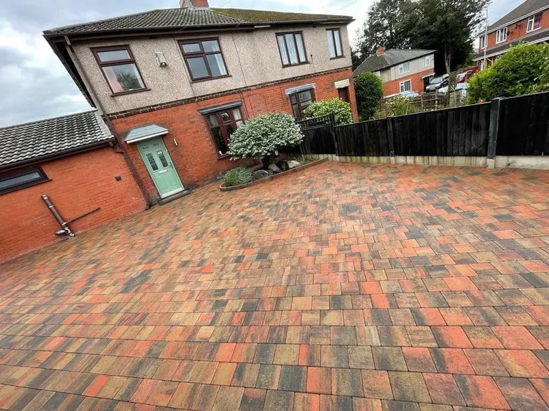 Long brindle herringbone driveway between a new timber fence with decorative wrought-iron gates and a tall hedge, finished with a charcoal block perimeter border.