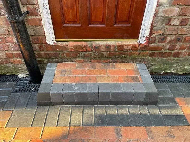 Large front area paved in red, charcoal and buff blocks laid in stretcher bond across a semi-detached house, with a circular raised planter set into the surface.