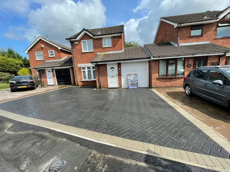 Downward-sloping brindle herringbone driveway with a double charcoal border running alongside a new vertical timber fence with concrete posts and gravel boards.
