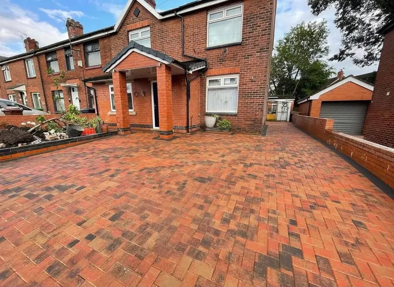 Charcoal herringbone driveway in front of a red brick house and white garage, framed by two rows of cream blocks where it meets the road and neighbouring boundaries.