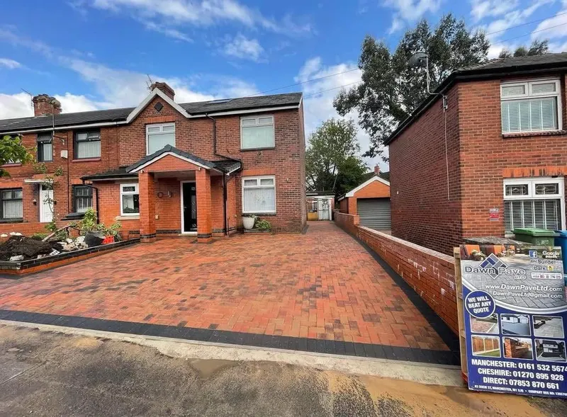 Charcoal herringbone paving before a white garage and front door, separated from a light block perimeter by a black linear drainage channel along the house wall.