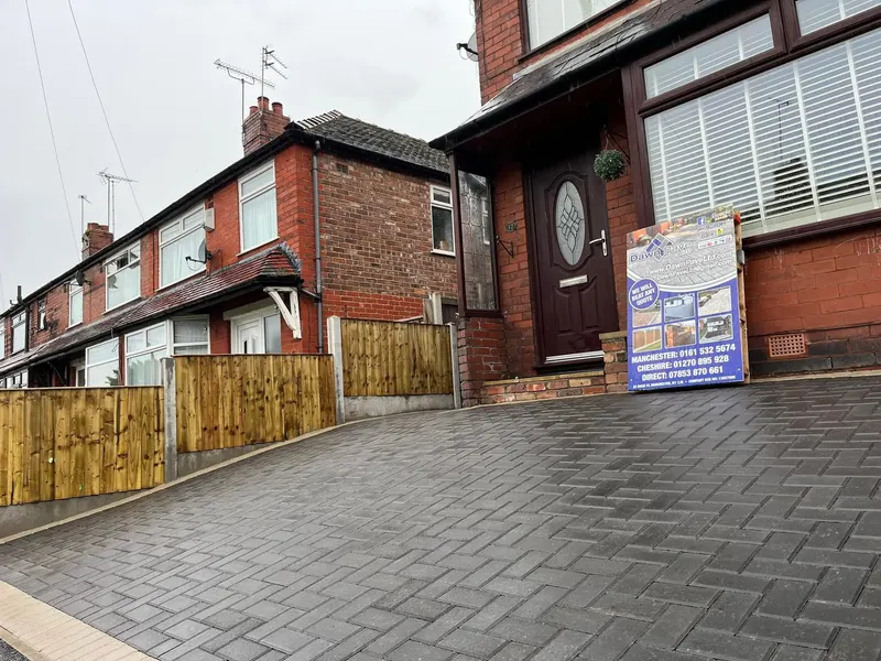 Full-width charcoal herringbone driveway on a white rendered house with solar panels, finished with a single row of tan blocks along the road edge.