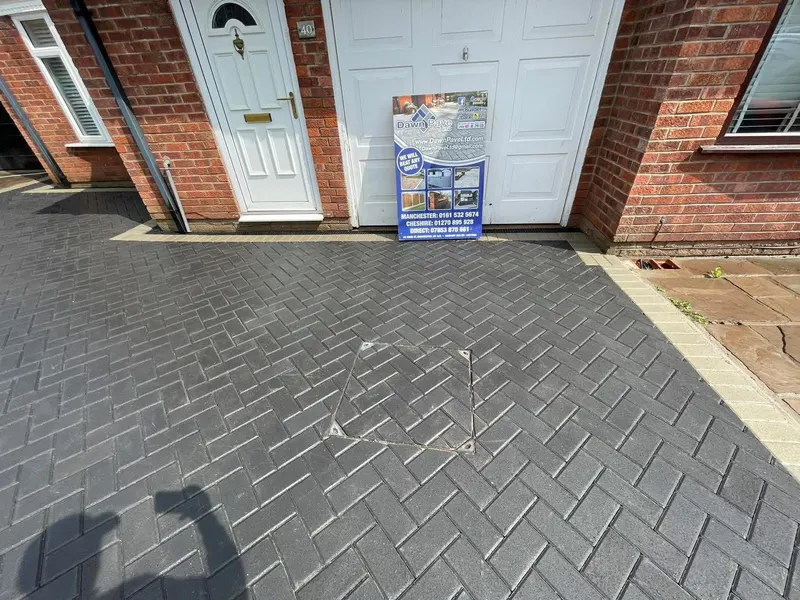 Charcoal herringbone driveway rising from the street to a red brick semi-detached house, with a light grey block border along the lower edge beside a new wooden fence.