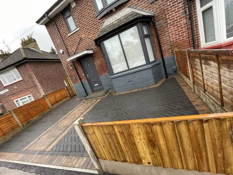 Charcoal herringbone driveway with buff border before a red brick house, including a recessed manhole infilled with matching herringbone blocks beside older slab paving.