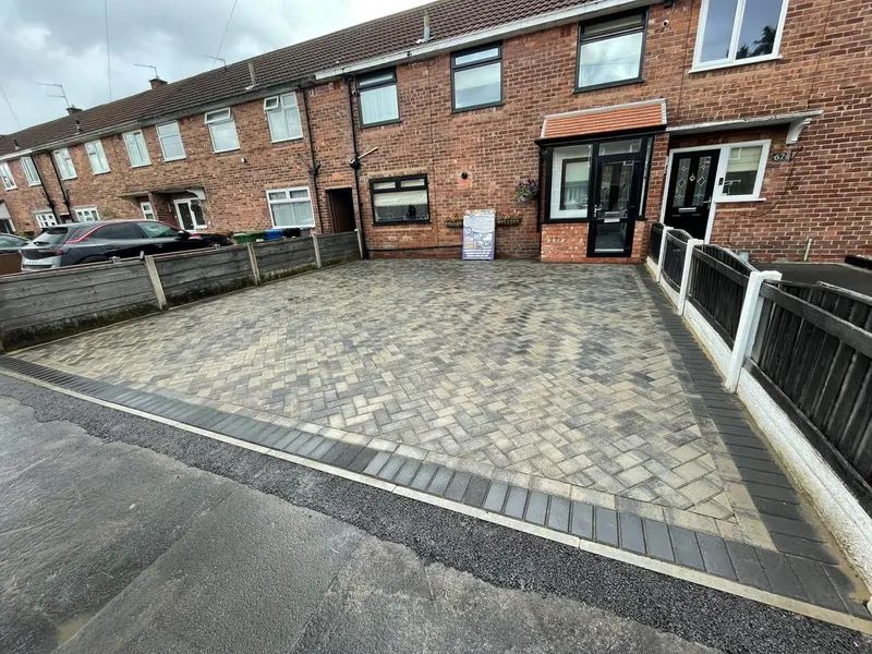 Light buff herringbone paving along a yellow-brick house with a soldier-course curved edge meeting tarmac, bounded by a red brick wall and wooden side gate.