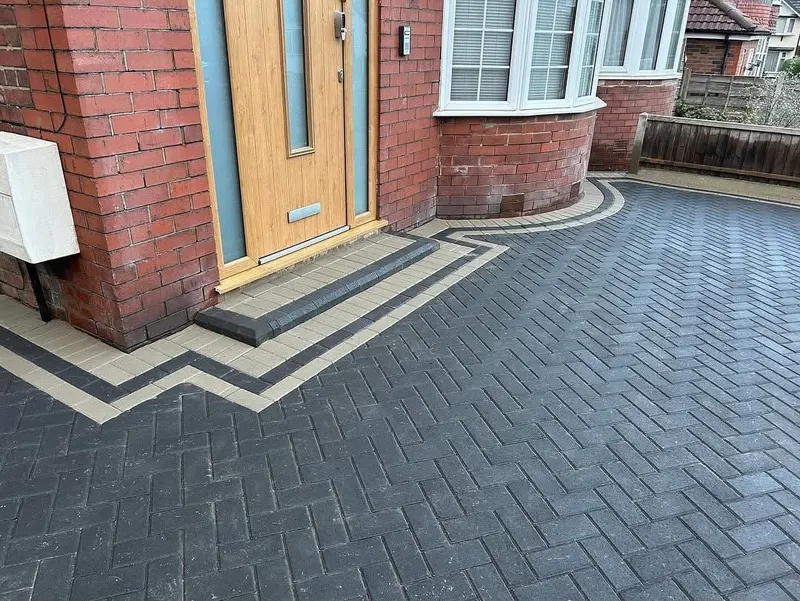Large charcoal herringbone driveway with charcoal header border, a recessed manhole set in the pattern, and entrance steps lined with timber sleepers filled with block paving.