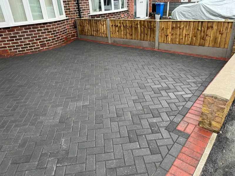 Charcoal herringbone driveway sweeping around a red brick bay window, with a three-course decorative border in cream and charcoal blocks and a small kerbed doorstep.