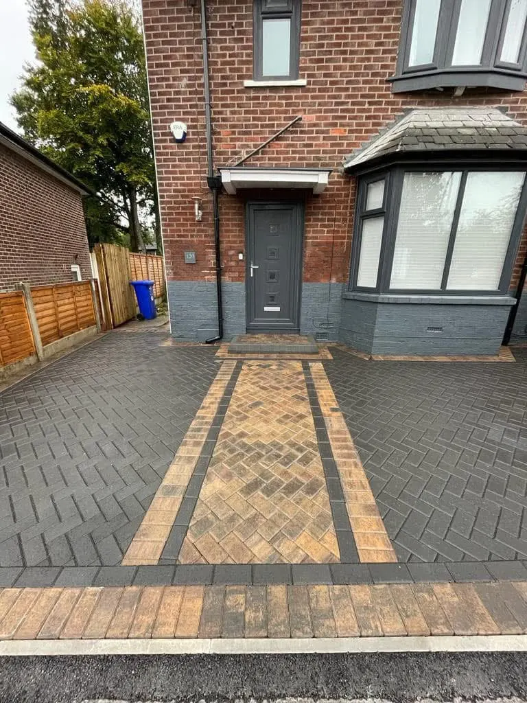 Charcoal herringbone driveway curving along a red brick house and timber fence, finished with a red block soldier border and a stone block boundary wall.