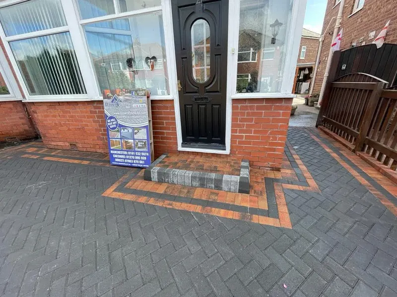 Charcoal herringbone paving beside a red brick wall and white door, with soldier-course edging, a channel drain at the threshold, and a manhole infilled to match the pattern.