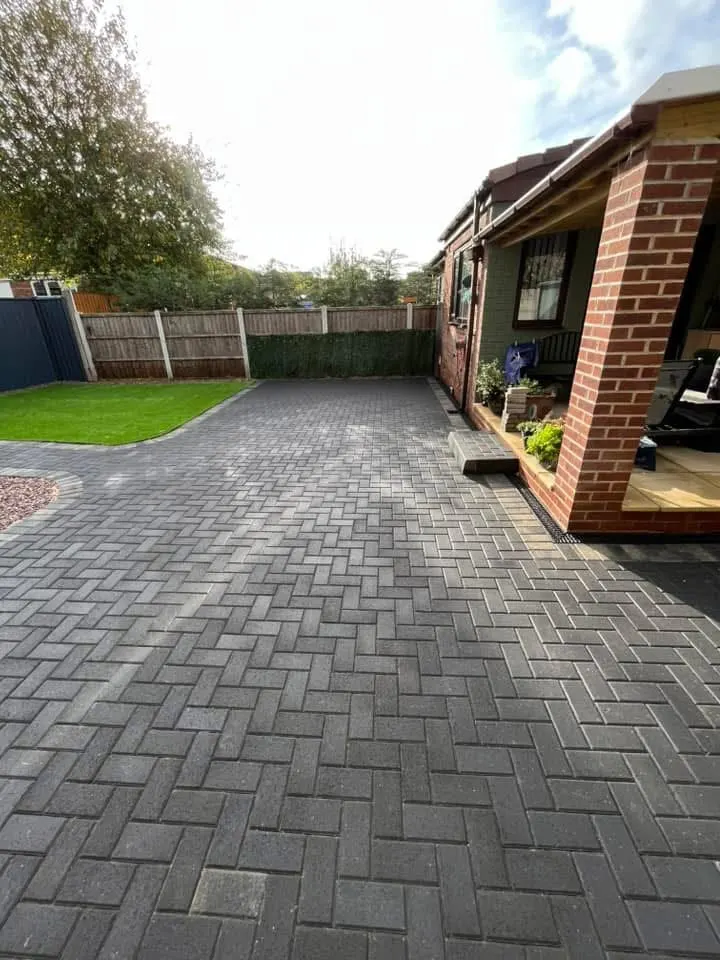 Straight red terracotta herringbone driveway with charcoal double border at the kerb, running between new wooden panel fences toward a red brick semi-detached house.