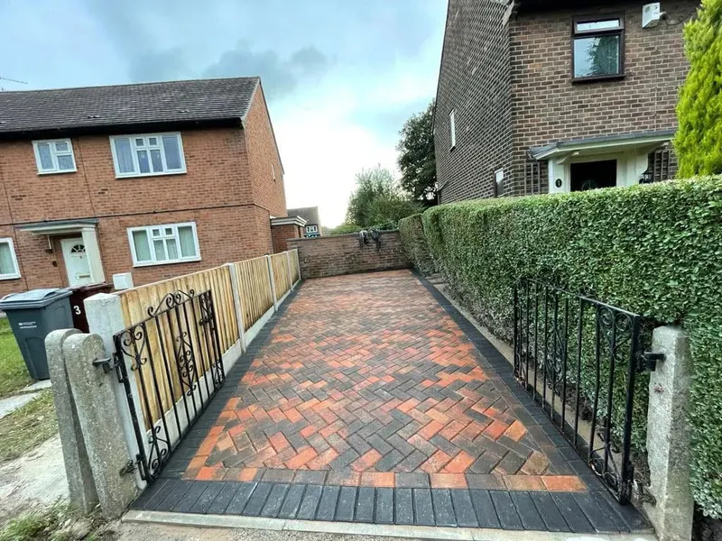 Sandy buff herringbone paving along a yellow-brick house wall with soldier-course edging on a curved transition to asphalt, beside a red brick boundary wall.