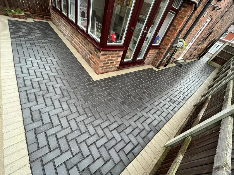 Long brindle herringbone driveway with charcoal perimeter border, leading from open wrought-iron gates between a trimmed hedge and a light timber fence.