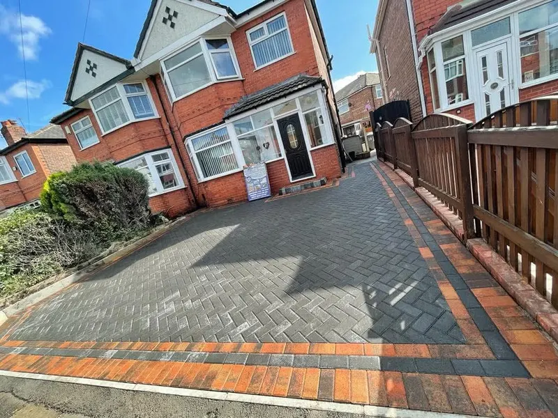 Large charcoal herringbone driveway before a detached red brick house, edged with tan blocks and an outer charcoal soldier course beside timber sleeper planting beds.