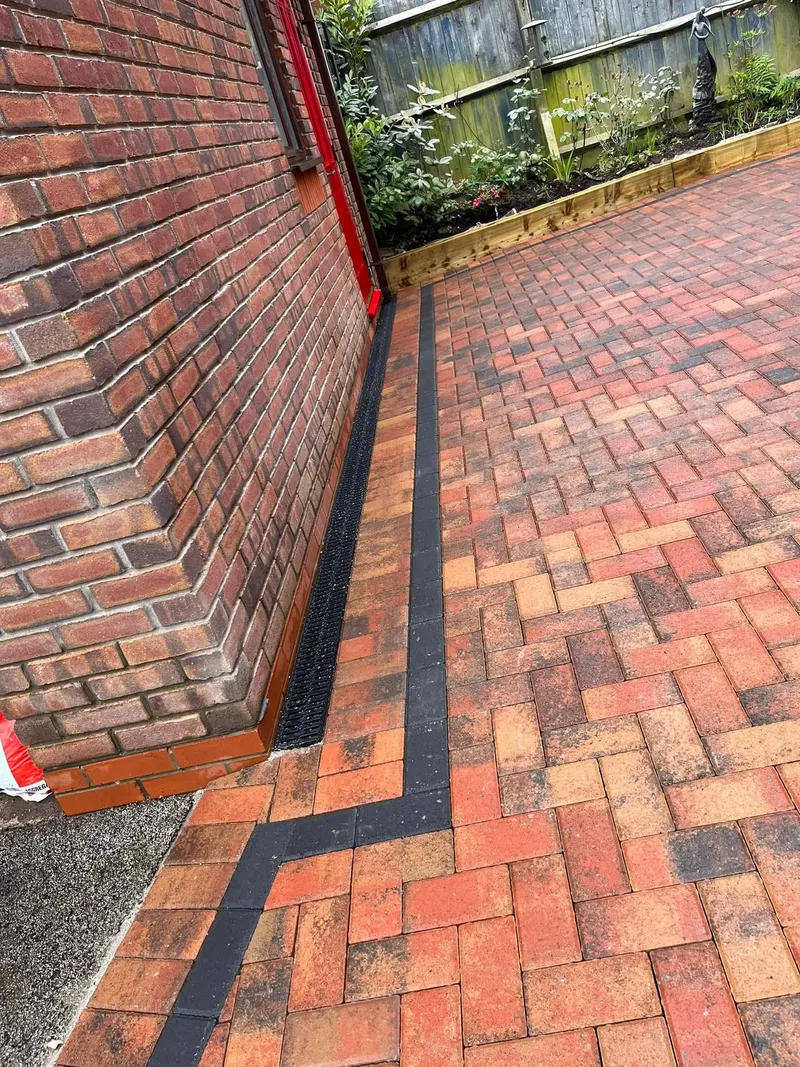 Charcoal herringbone driveway between brick entrance pillars up to grey door and garage on a red brick house, surrounded by a buff block border and small front lawn.