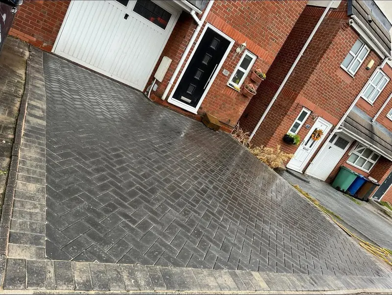 Red multi-tonal herringbone paving turning the corner of a brick house, with charcoal soldier border and a black channel drain fixed along the wall beside a timber planter.