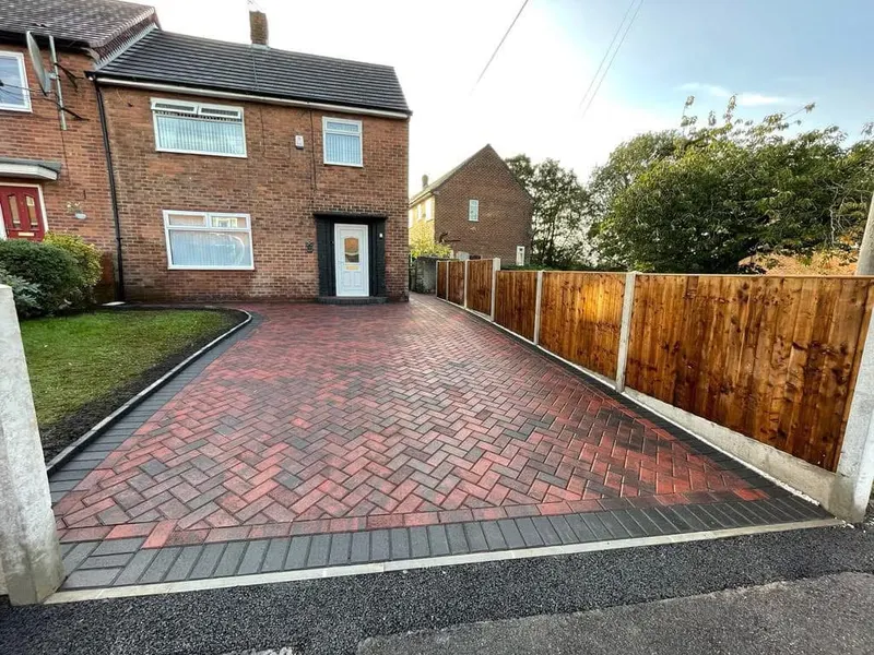 Brindle herringbone driveway with a large charcoal geometric inset of interlocking squares near the garage, charcoal edging and a channel drain along the brick wall.