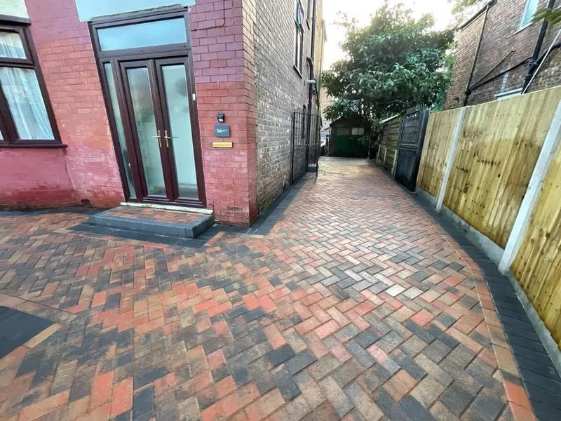 Brindle herringbone driveway with charcoal double border at the kerb, curving beside a small lawn on one side and a new timber fence on the other.