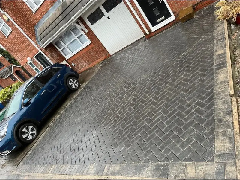 Extended multicolour herringbone driveway between dark panel fences, leading to a semi-detached house with red brick ground floor and pebbledash upper storey.