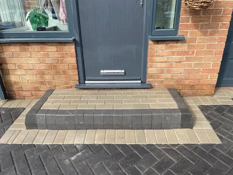 Long brindle mixed-size block path between a red brick wall and dark timber fence, with charcoal soldier edging, channel drain and a curved planter border at the far end.