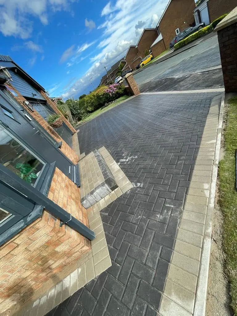 Charcoal herringbone driveway with a central tan herringbone walkway and brown soldier border bands, between timber fences on a grey-accented red brick house.