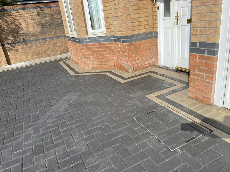 Close-up of charcoal herringbone paving beside red brickwork, with soldier-course edging and a recessed tray infilled so the manhole cover matches the surrounding pattern.