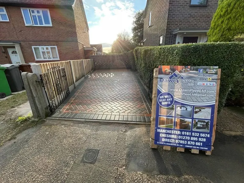 Broad charcoal herringbone driveway with dark soldier border and pale stone edging, spanning a red brick house front garden to the tarmac road under cloudy skies.