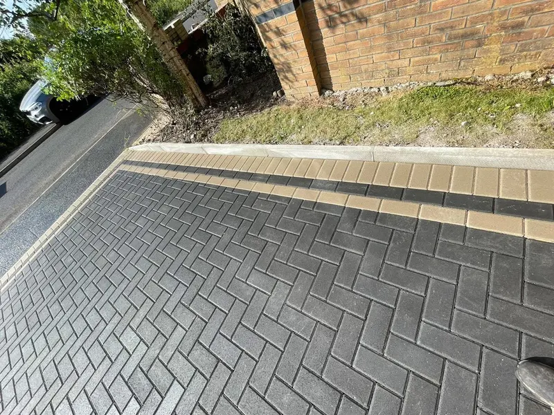 Brindle and charcoal herringbone parking area between a timber fence with small gate and a tall hedge, backed by a low brick wall on a sunny residential plot.