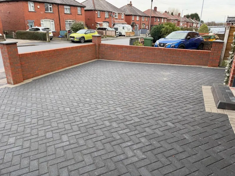 Charcoal herringbone driveway detail showing buff-charcoal-buff triple border courses against a red brick wall, with kerb edging and street visible beyond.