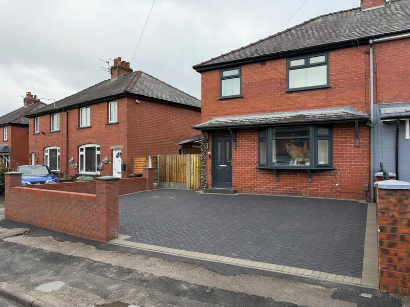 Charcoal herringbone driveway on a red brick semi with grey window frames, edged in light grey header blocks and bounded by a low matching brick wall on one side.