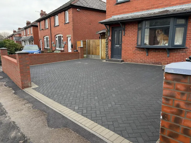 Charcoal herringbone driveway with buff front edging before a red brick semi-detached house, enclosed on the left by square-pier brick wall with grey door and windows.